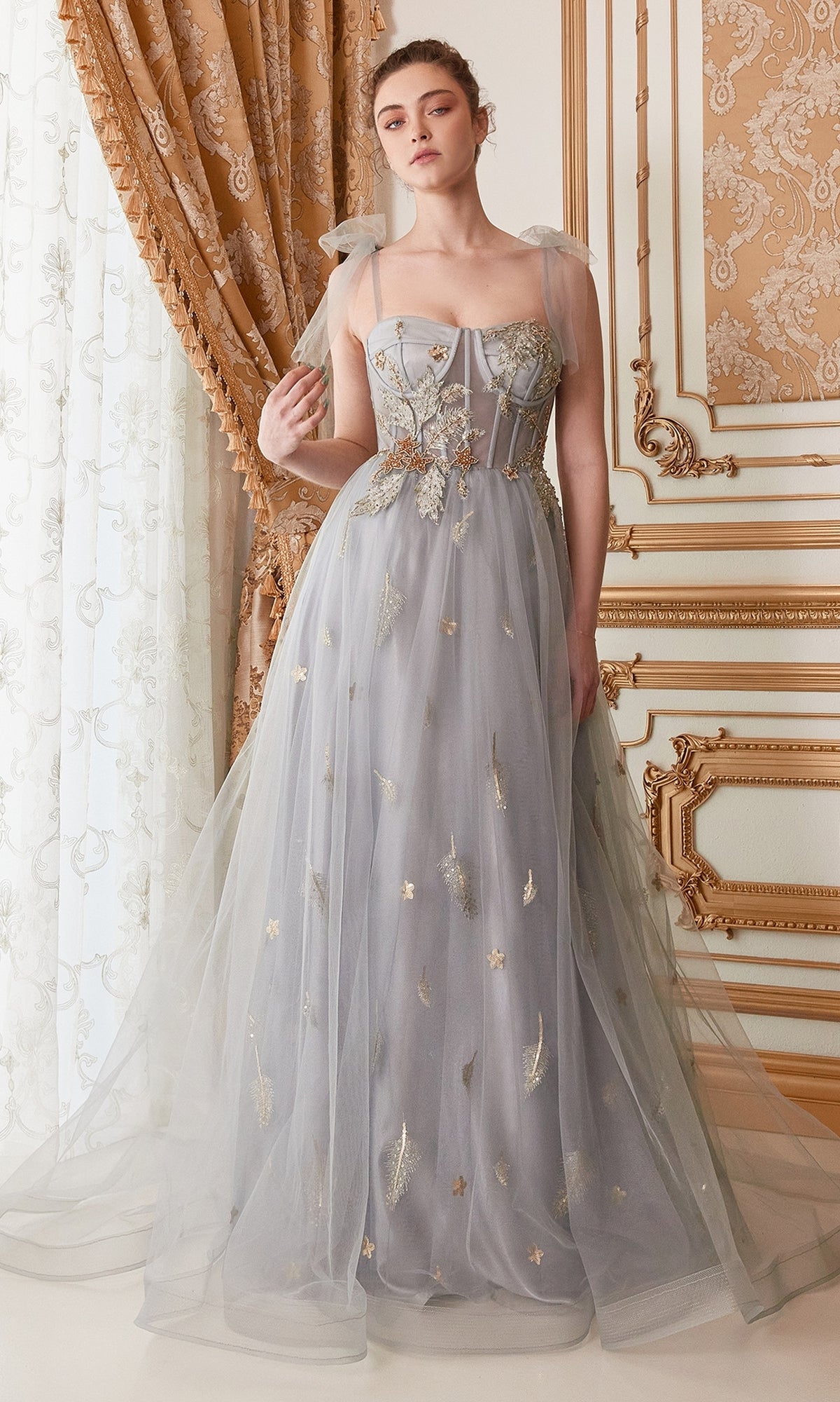 A woman stands indoors in front of ornate curtains wearing the Shoulder-Bow Long Corset Prom Ball Gown A0824, a light blue tulle dress with gold embroidery, sheer shoulder bows, and a fitted corset bodice.
